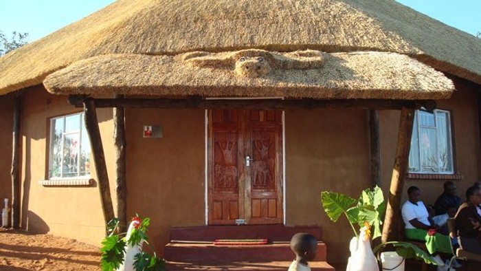 et billede af en hytte/skole i Zambia i Afrika, et barn udenfor og mennesker sidder op af skolen