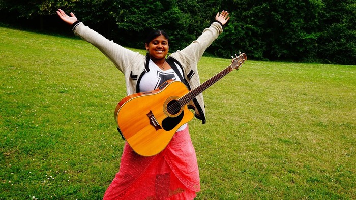 Ankita står på græsplæne med sin guitar på maven og begge hænder over hovedet.