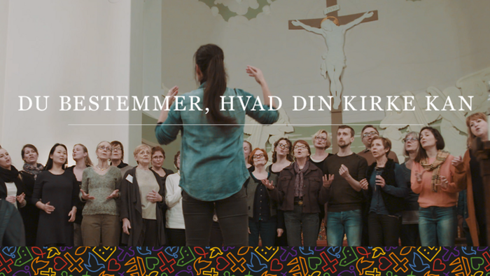 Et kor synger sammen i en kirke med en dirigent, der leder, under et kors og tekst på dansk.