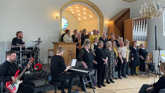 En gruppe synger og spiller instrumenter i en kirke, ledet af en korleder ved et podie.