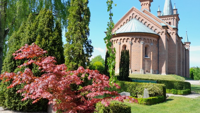 En smuk kirke med røde mursten og spisse tårne, omgivet af grønne træer og buske.