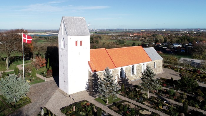 En lille kirke med tårn og rødt tag, beliggende i en landsby med en dansk flag.