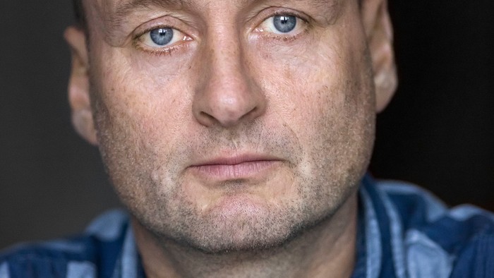 Et nærbillede af en skaldet mand iført en blåstribet skjorte mod en mørk baggrund.