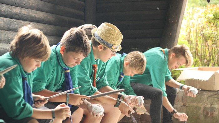 Eine Gruppe von Kindern in grünen Shirts beteiligt sich an einer Outdoor-Aktivität und geht vorsichtig mit kleinen Gegenständen um, die sie mit Stöcken handhaben.
