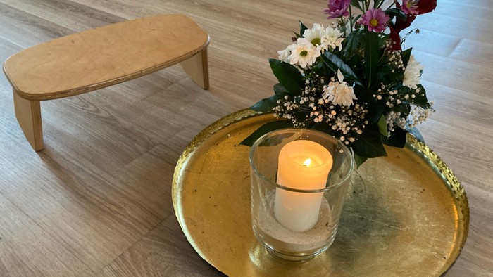 Eine brennende Kerze und eine Vase mit Blumen auf einem goldenen Tablett neben einer Holzbank.