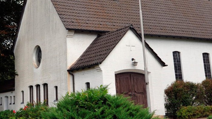 Weiße Kirche mit hohem Turm und braunem Dach, umgeben von Bäumen und Büschen