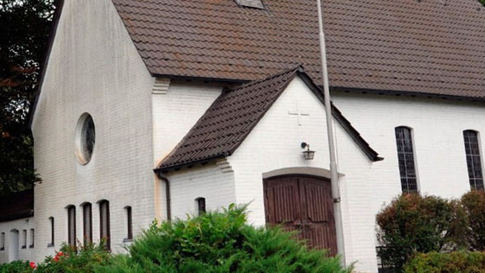 Weiße Kirche mit hohem Turm und braunem Dach, umgeben von Bäumen und Büschen