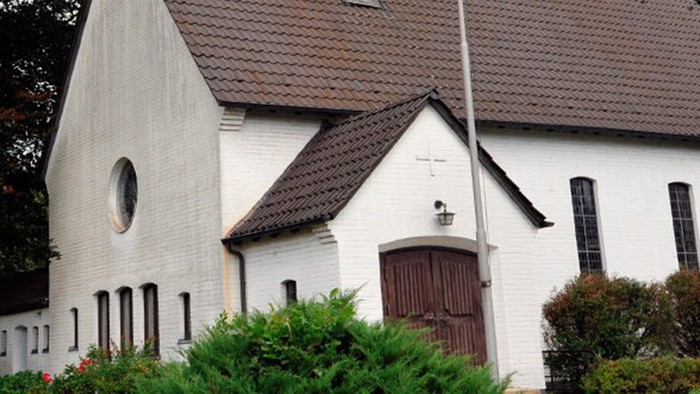 Weiße Kirche mit hohem Turm und braunem Dach, umgeben von Bäumen und Büschen