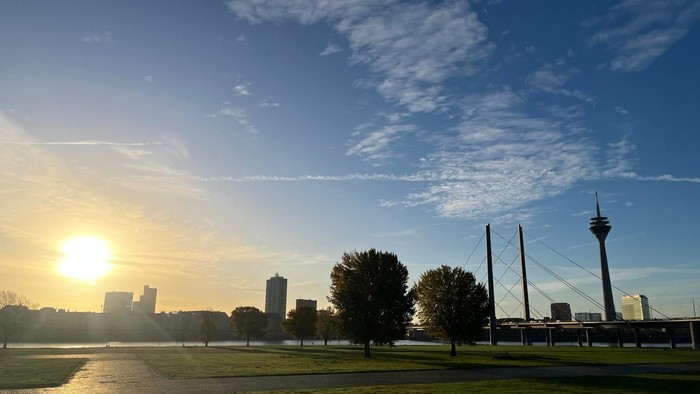 Sonnenuntergang über einer Stadt-Skyline mit einem hohen Turm, einem grünen Park und einem ruhigen Fluss, der das Licht reflektiert.