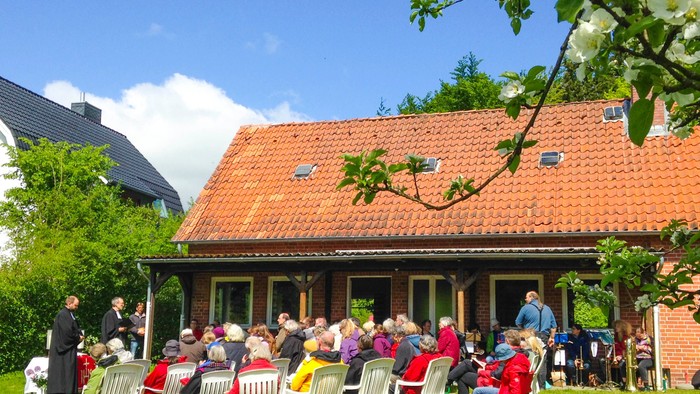 Eine Gruppe von Menschen sitzt auf Stühlen vor einem Haus mit rotem Dach.