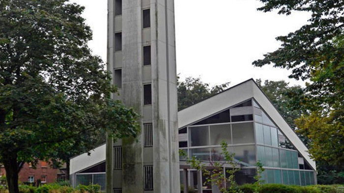 Das Bild zeigt eine moderne Kirche mit hohem Turm und Glasfront, umgeben von Bäumen.