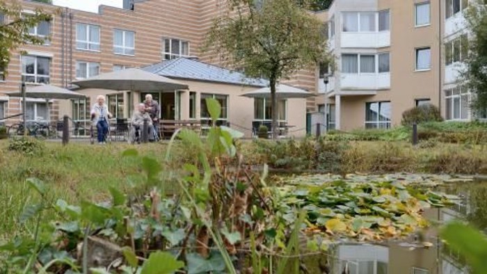 Im Bild ist ein ruhiger Innenhof mit einem Teich und einem Pavillon zu sehen, umgeben von Wohngebäuden.