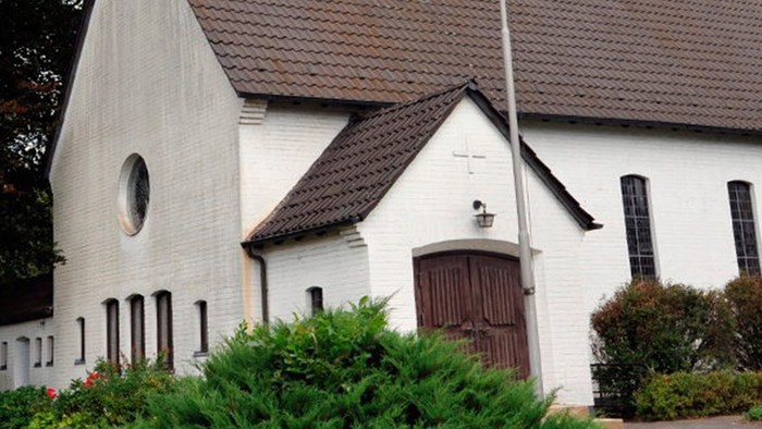 Weiße Kirche mit hohem Turm und braunem Dach, umgeben von Bäumen und Büschen