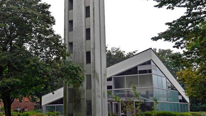 Das Bild zeigt eine moderne Kirche mit hohem Turm und Glasfront, umgeben von Bäumen.