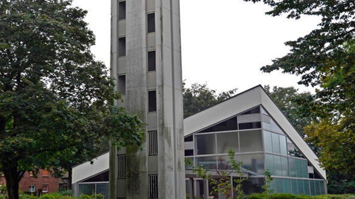Das Bild zeigt eine moderne Kirche mit hohem Turm und Glasfront, umgeben von Bäumen.