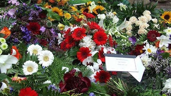 Farverigt blomsterarrangement ved et udendørs mindesmærke med en plade mellem livlige blomster.