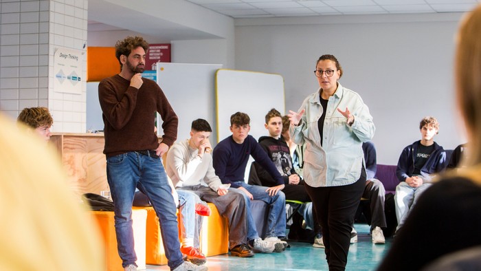Zwei Moderator:innen leiten eine interaktive Sitzung mit einer Gruppe sitzender Teilnehmer:innen in einem Klassenzimmer.