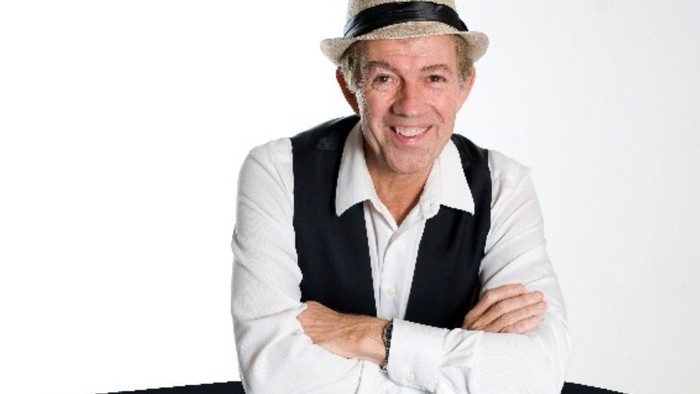 foto af Stig Nørregaard, smilende herre med lys hat med sort hattebånd, hvid skjorte og sort vest, læner sig smilende forover med armene over kors / på albuerne på et sort bord