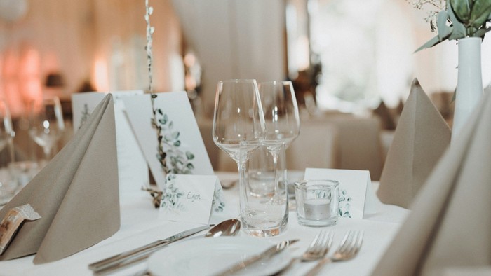 Elegant opstillet bord med vinglas, bestik, servietter og diskrete blomsterplaceringer til en formel begivenhed.