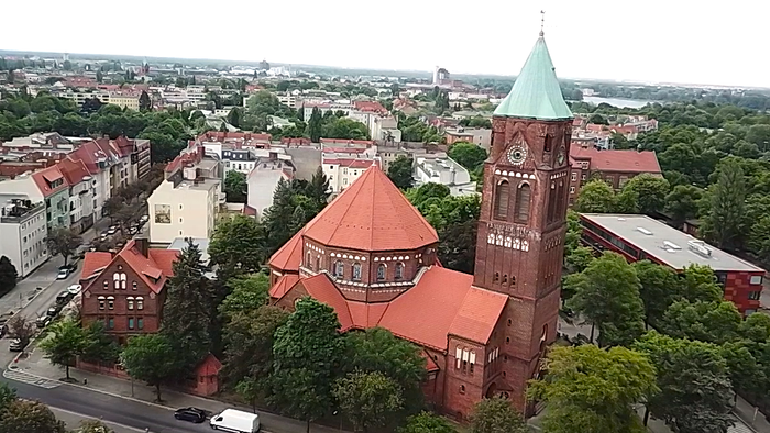 Luftaufnahme einer historischen Backsteinkirche mit hohem grünem Turm in einer grünen, städtischen Umgebung.