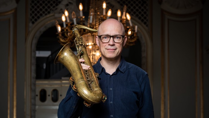 En skaldet mand med briller holder en messing-saxofon i et prægtigt, svagt oplyst rum.