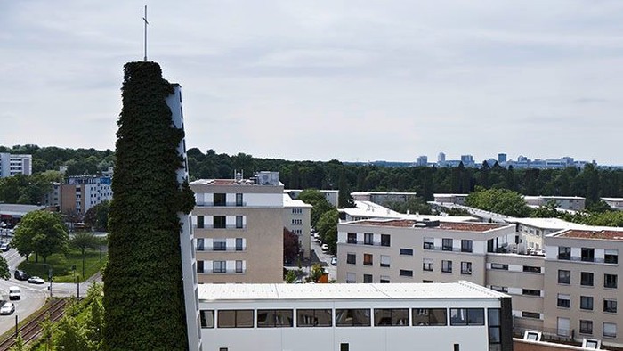 Städtische Szene mit einem hohen Schornstein neben modernen Gebäuden und Grünflächen unter einem bewölkten Himmel.