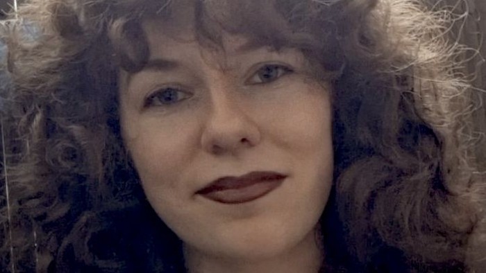 Eine Person mit lockigem Haar und neutralem Gesichtsausdruck in einer sanft beleuchteten Innenraumeinstellung.