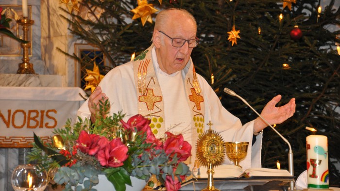 Ein Priester in weißer Soutane hält eine Predigt an einem Altar mit Weihnachtsdekoration.