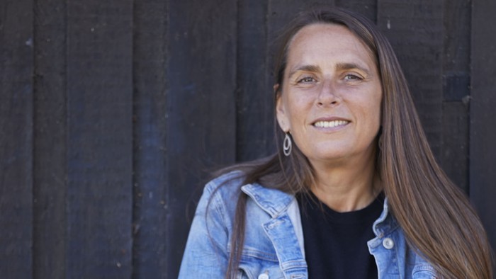 En selvsikker kvinde i en denimjakke står med armene over kors foran en mørk trævæg.