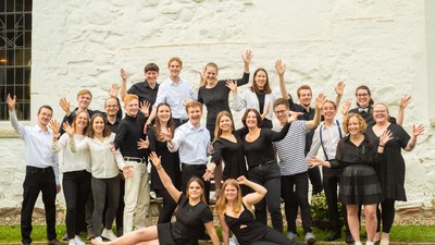 Eine Gruppe junger Menschen posiert vor einer weißen Steinmauer, viele halten den Peace-Zeichen