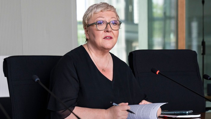 En kvinde med briller sidder ved et bord og skriver noter under det, der ser ud til at være et formelt møde eller en session.