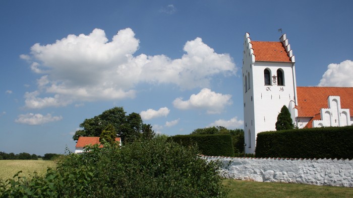 En hvid kirke med rødt tag og klokketårn ved en murstenet vegg og grønne buske
