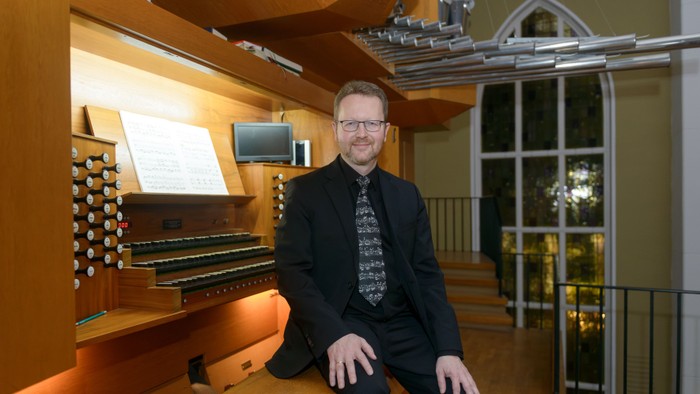 Ein Mann sitzt neben einer großen Orgel in einem hellen, eleganten Raum mit großen Fenstern.
