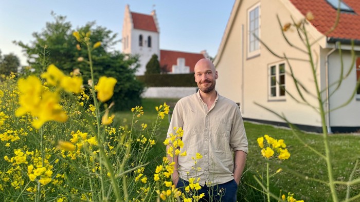 En mand står midt i levende gule blomster i en frodig have med et idyllisk hus i baggrunden.