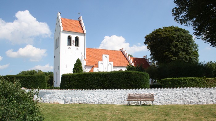 En hvid kirke med rødt tag og tårn, omgivet af en mur og buske, med en bænk foran