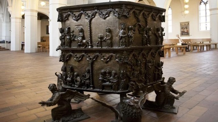 Bronzener Taufstein mit reliefartigen Figuren und Verzierungen, getragen von vier knienden Figuren.
