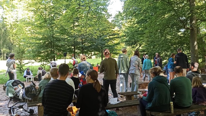 Eine Gruppe von Menschen versammelt sich im Freien in einem bewaldeten Gebiet, wahrscheinlich für ein Picknick oder eine Gemeinschaftsveranstaltung.