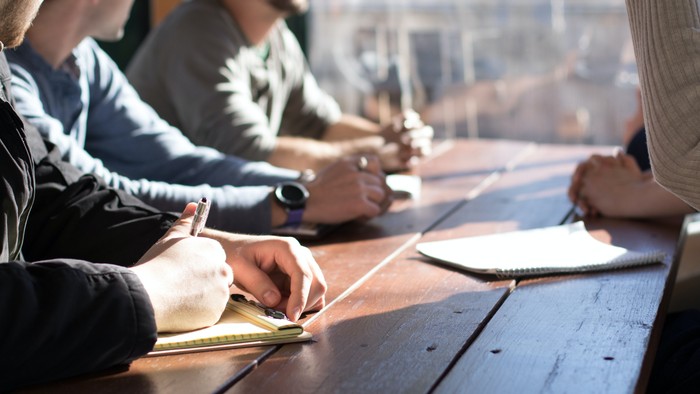 Männer sitzen an einem Tisch und arbeiten mit Notizbüchern und Kaffeetassen.