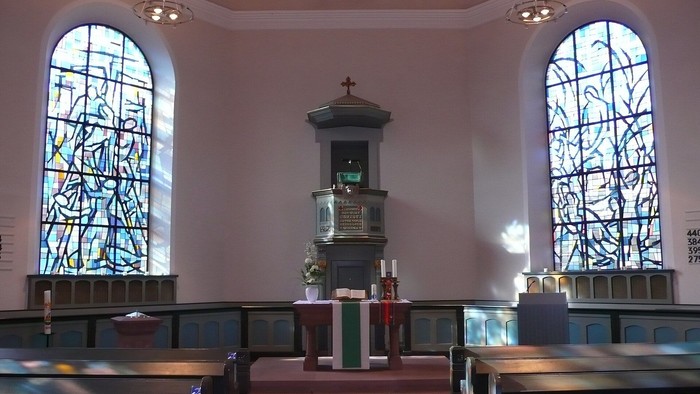 Ein ruhiger Kircheninnenraum mit Buntglasfenstern, hölzernen Kirchenbänken und einem zentralen Altarbereich.