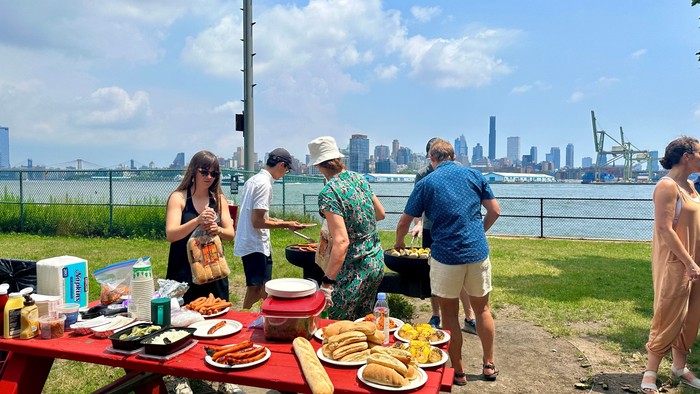 Mennesker, der nyder en picnic ved floden med grillet mad og byens skyline i baggrunden.