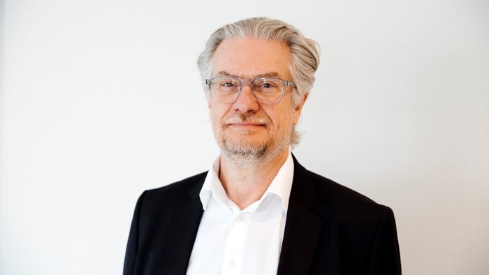 En mand med briller og formelt tøj poser foran en ensfarvet baggrund med et neutralt udtryk.