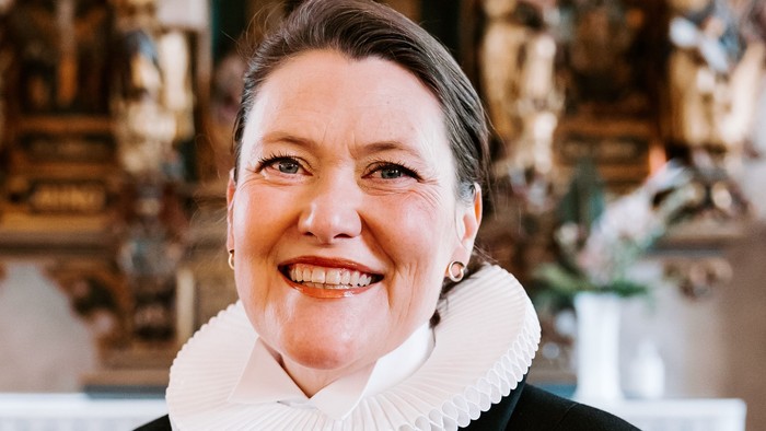 En kvinde i sort jakke med hvid krage står foran en prægtig altertavle.