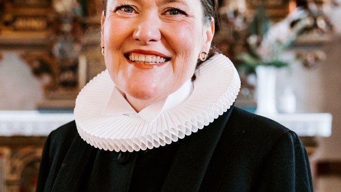 En kvinde i sort jakke med hvid krage står foran en prægtig altertavle.