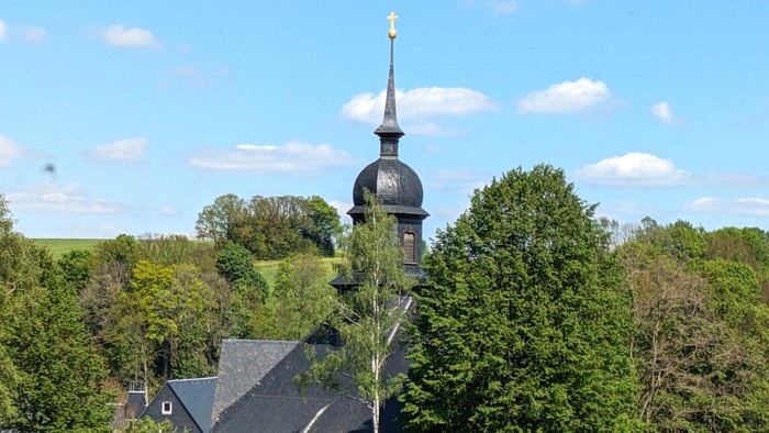Kirchturm mit Kreuz auf grünem Hügel, umgeben von Bäumen und Häusern