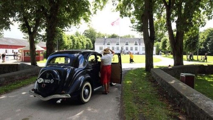 Gammel bil med en kvinde i tidstypisk kjole, der læsser bagage ved en historisk bygning.