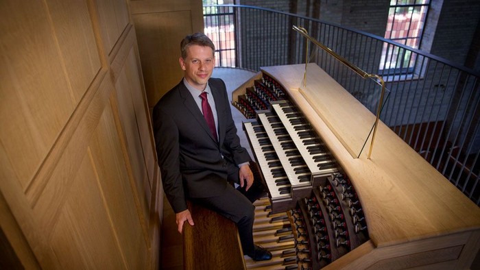 En mand i formelt tøj sidder ved siden af et stort pibeorgel i en kirke.