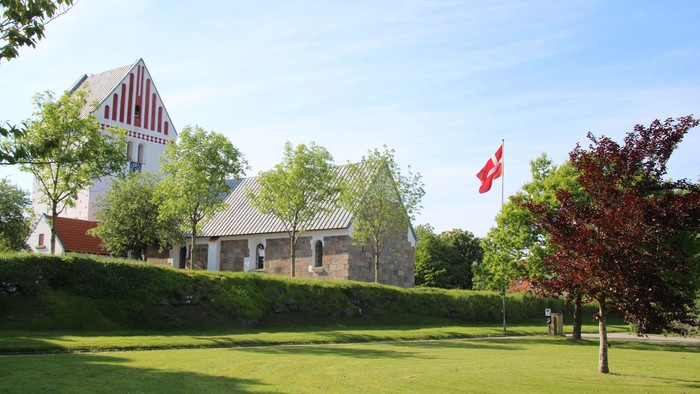 Historisk kirke med en flagstang, der viser et rød-hvidt nordisk korsflag i en frodig grøn omgivelse.