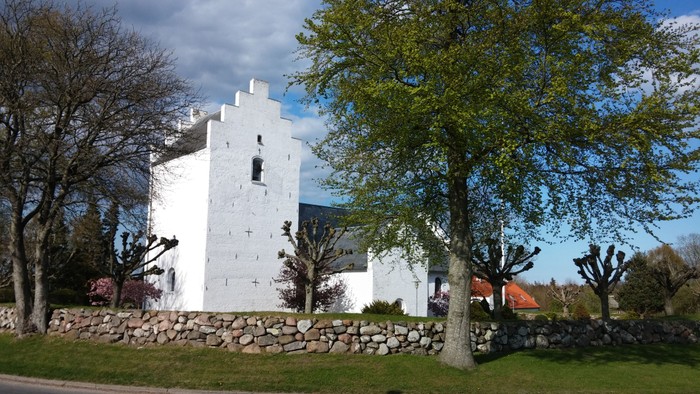 Et hvidt kirkebygning med tårn og murvej til, omgivet af træer og græs.