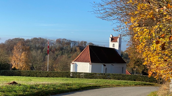 En lille hvid kirke med rødt tag og tårn ved en vej i en park med høstfarvede træer