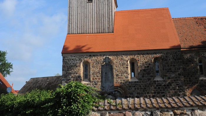 Kirchturm mit spitzem Dach und Uhr, umgeben von Steinmauer und Büschen